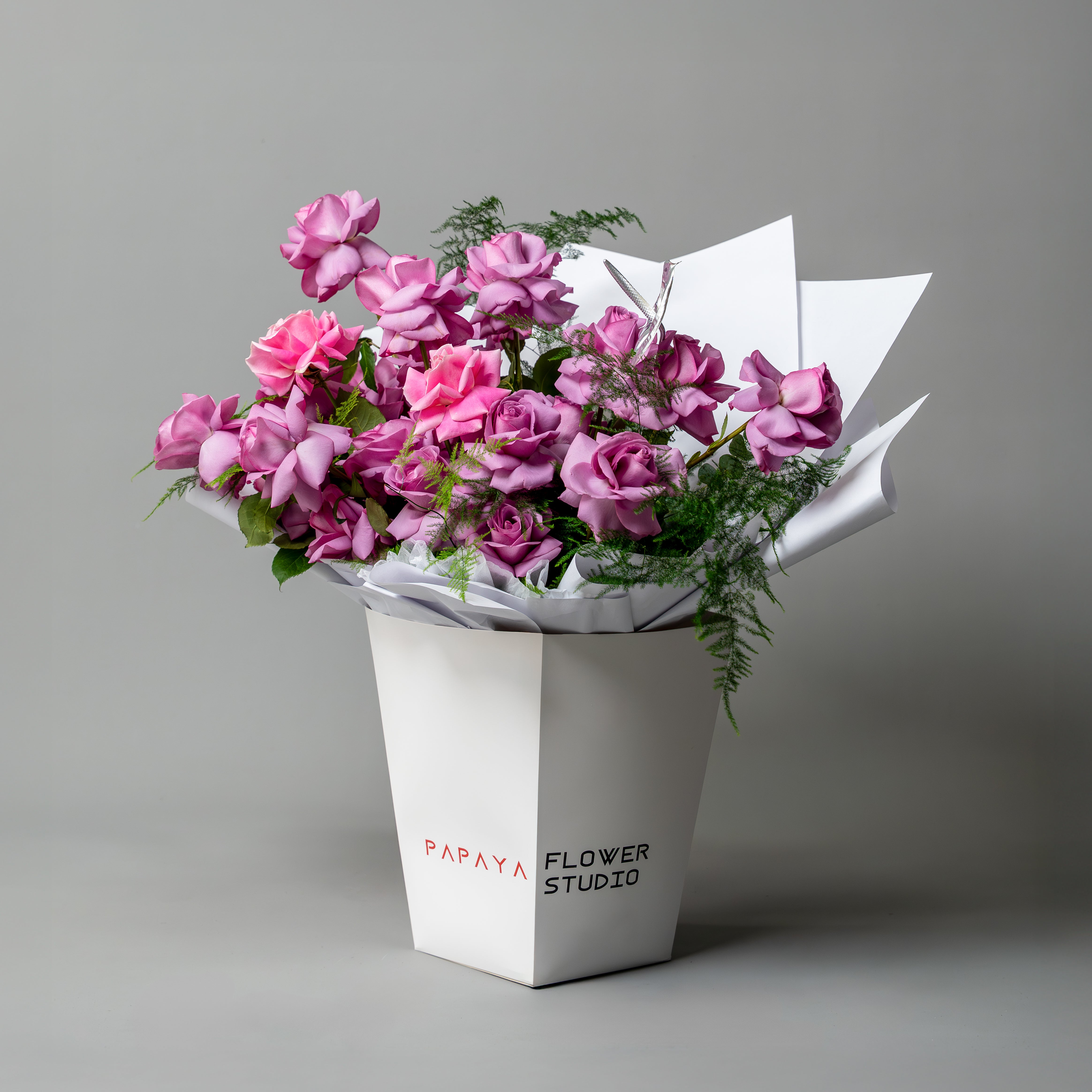 Vibrant bouquet of purple and pink reflexed roses with silver anthurium leaf in white branded carrier box delivery Dubai.