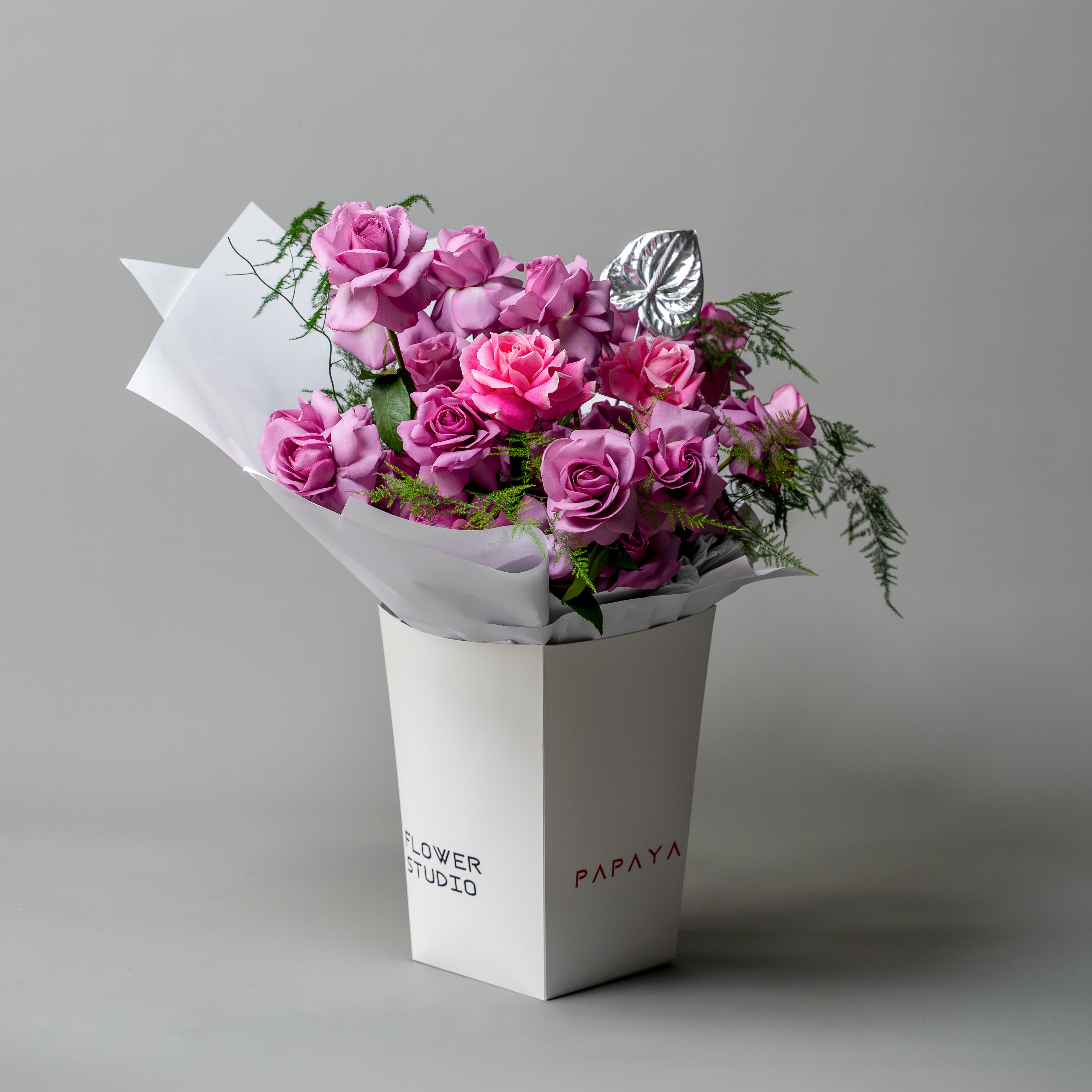 Vibrant bouquet of purple and pink reflexed roses with silver anthurium leaf in white branded carrier box delivery Dubai.