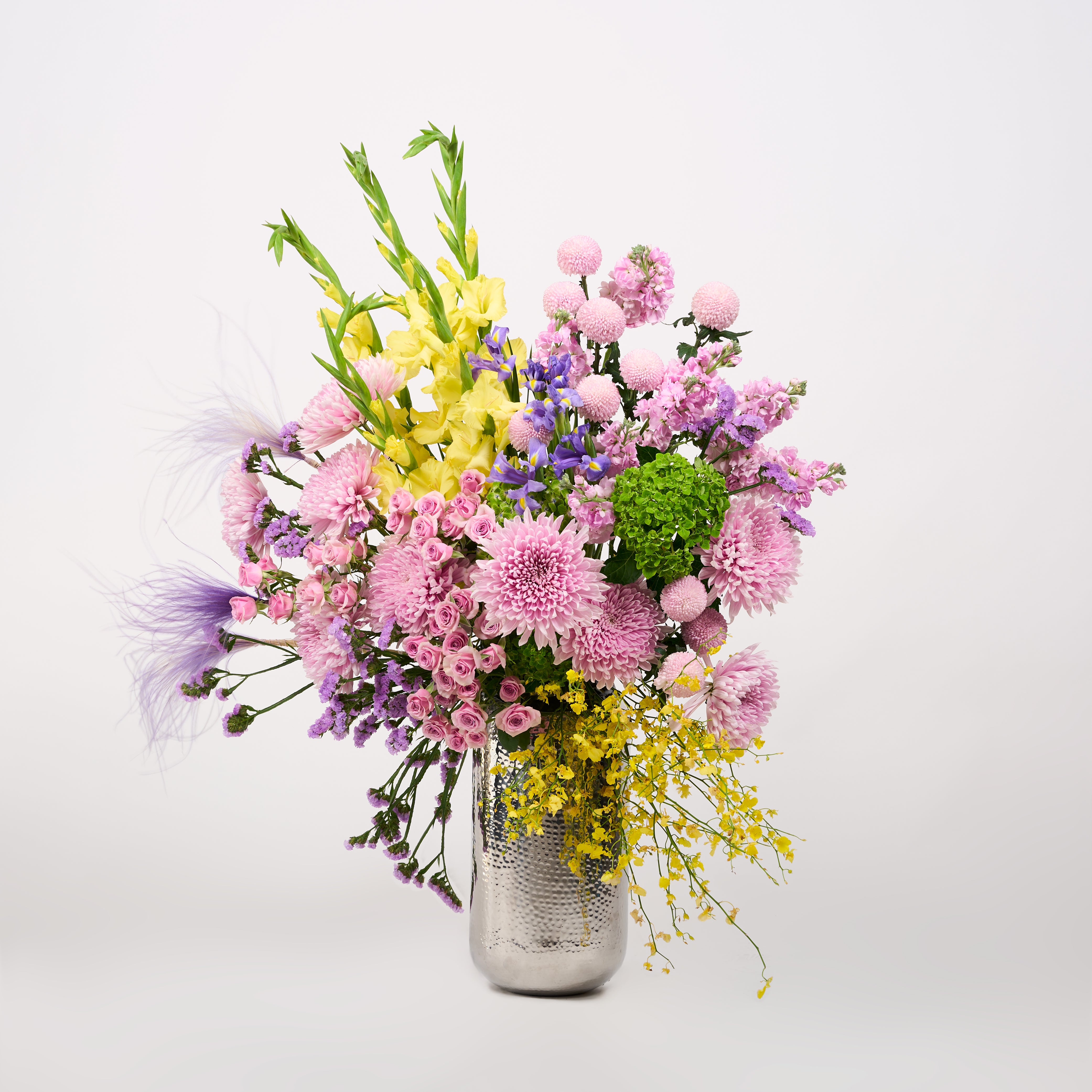 Pink, lilac and purple flowers with yellow blooms, green accents and feathery details in a textured silver-toned vase.