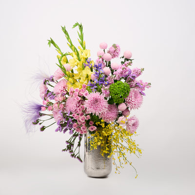 Pink, lilac and purple flowers with yellow blooms, green accents and feathery details in a textured silver-toned vase.
