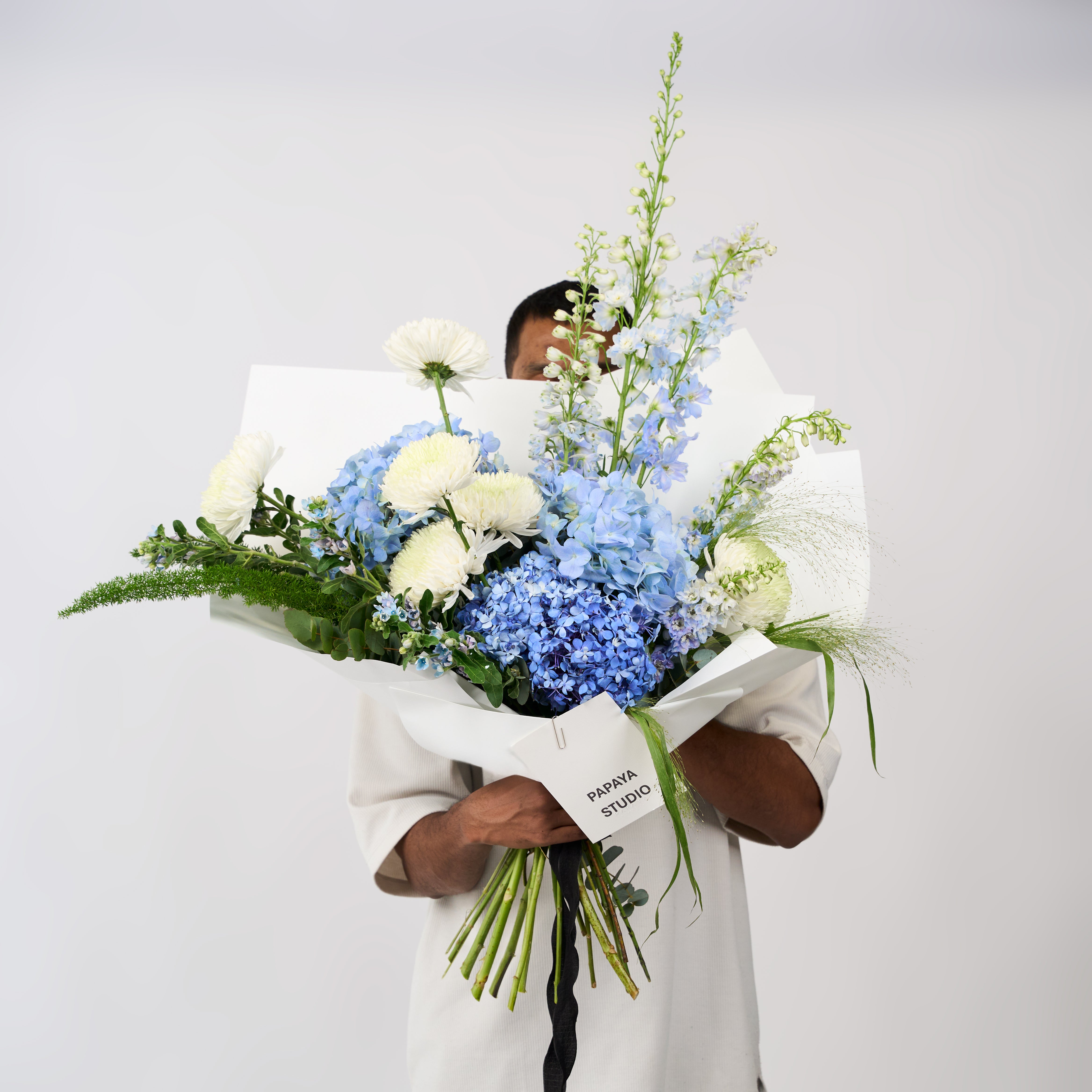 Blue hydrangea and creamy white flower bouquet with tall flowering stems, green foliage, ivory wrapping and a black ribbon.