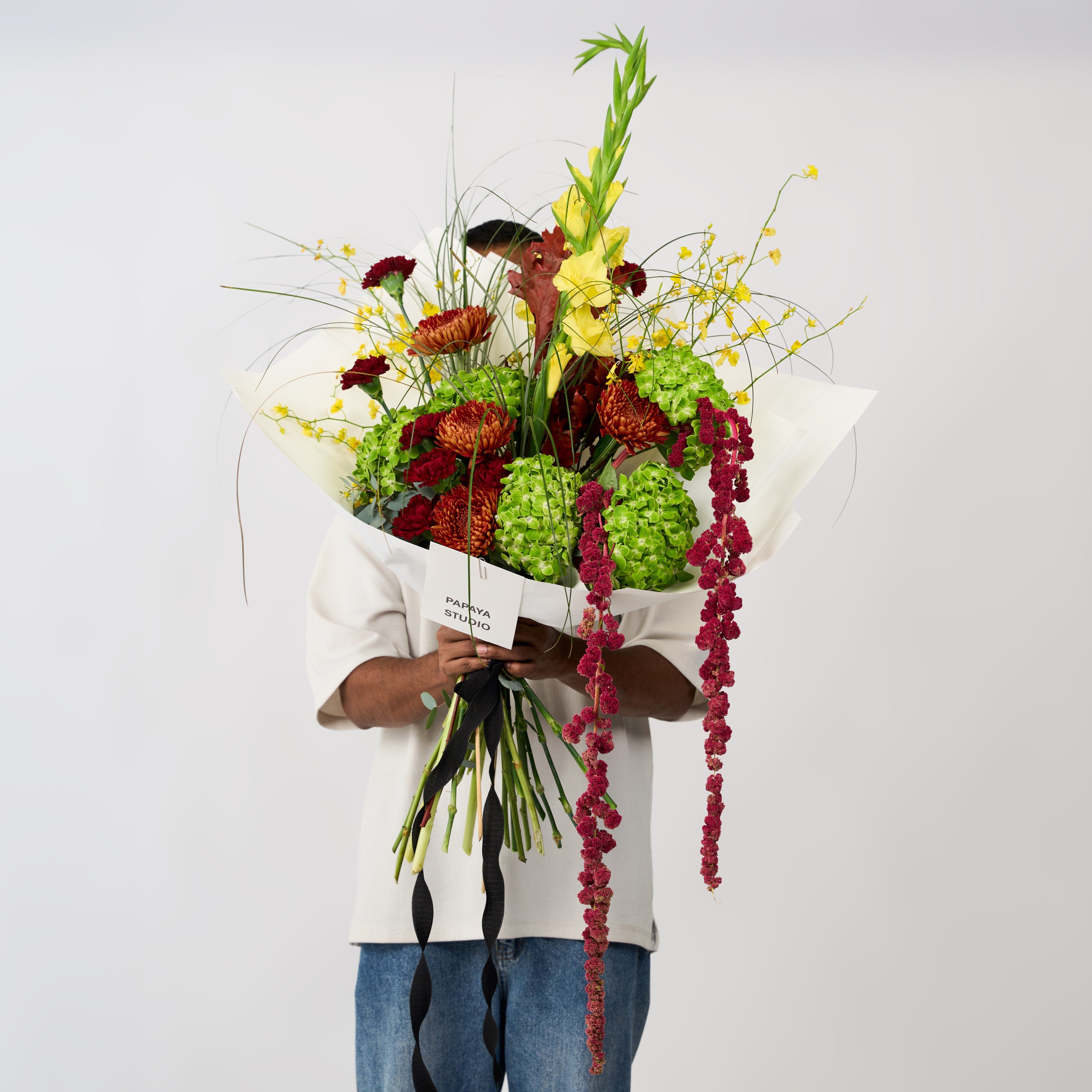 Green, yellow and copper flower bouquet with cascading burgundy blooms, tall stems, ivory wrapping and a black ribbon.