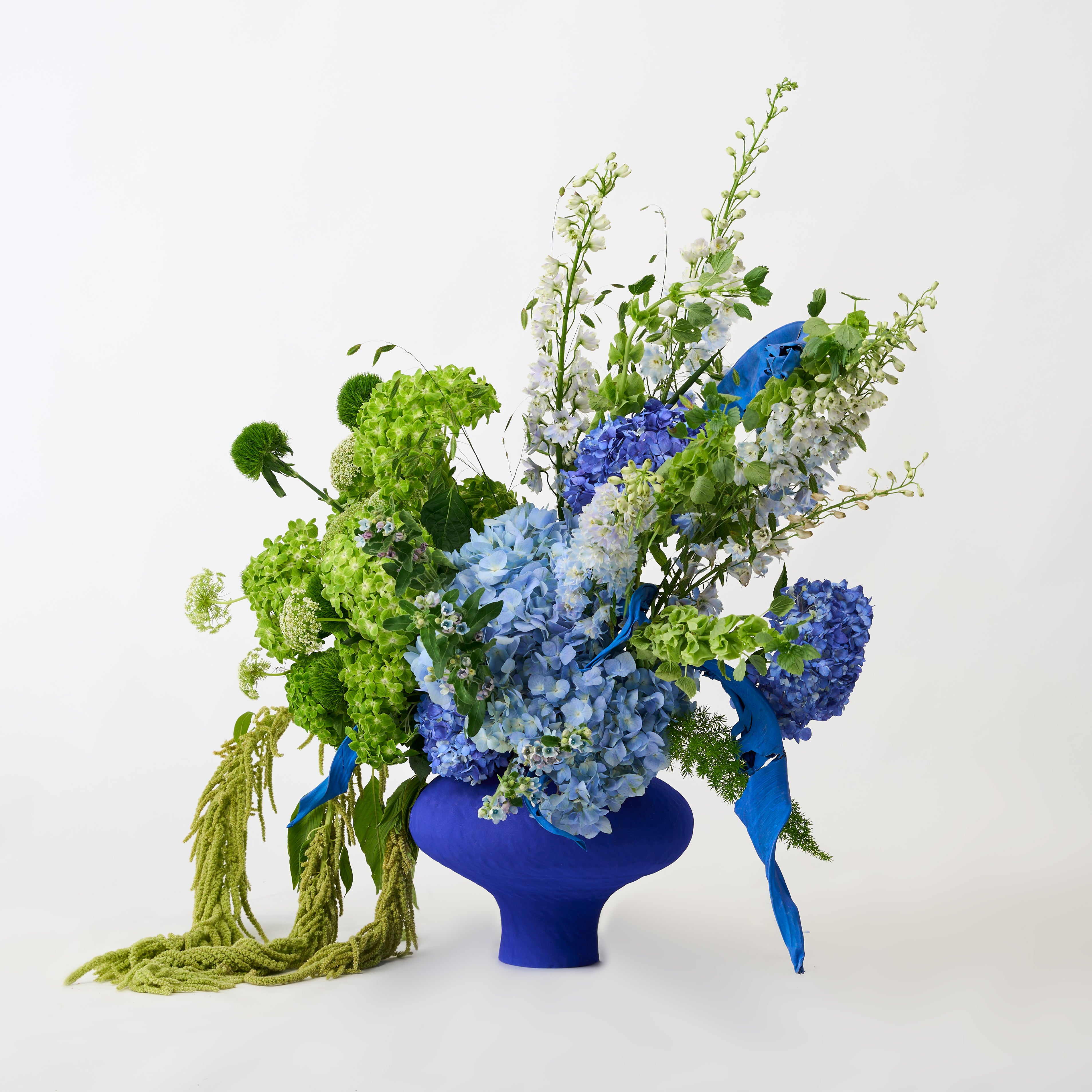 Blue and lime-green hydrangeas with pale flowers, cascading green stems and blue accents in a cobalt-blue vase.