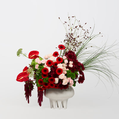Red and burgundy flowers with blush roses, green accents and sweeping grasses in a sculptural ivory vase.