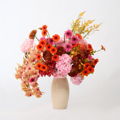 Pink hydrangeas, orange blooms and cascading golden orchids arranged in a tall, ribbed cream vase.