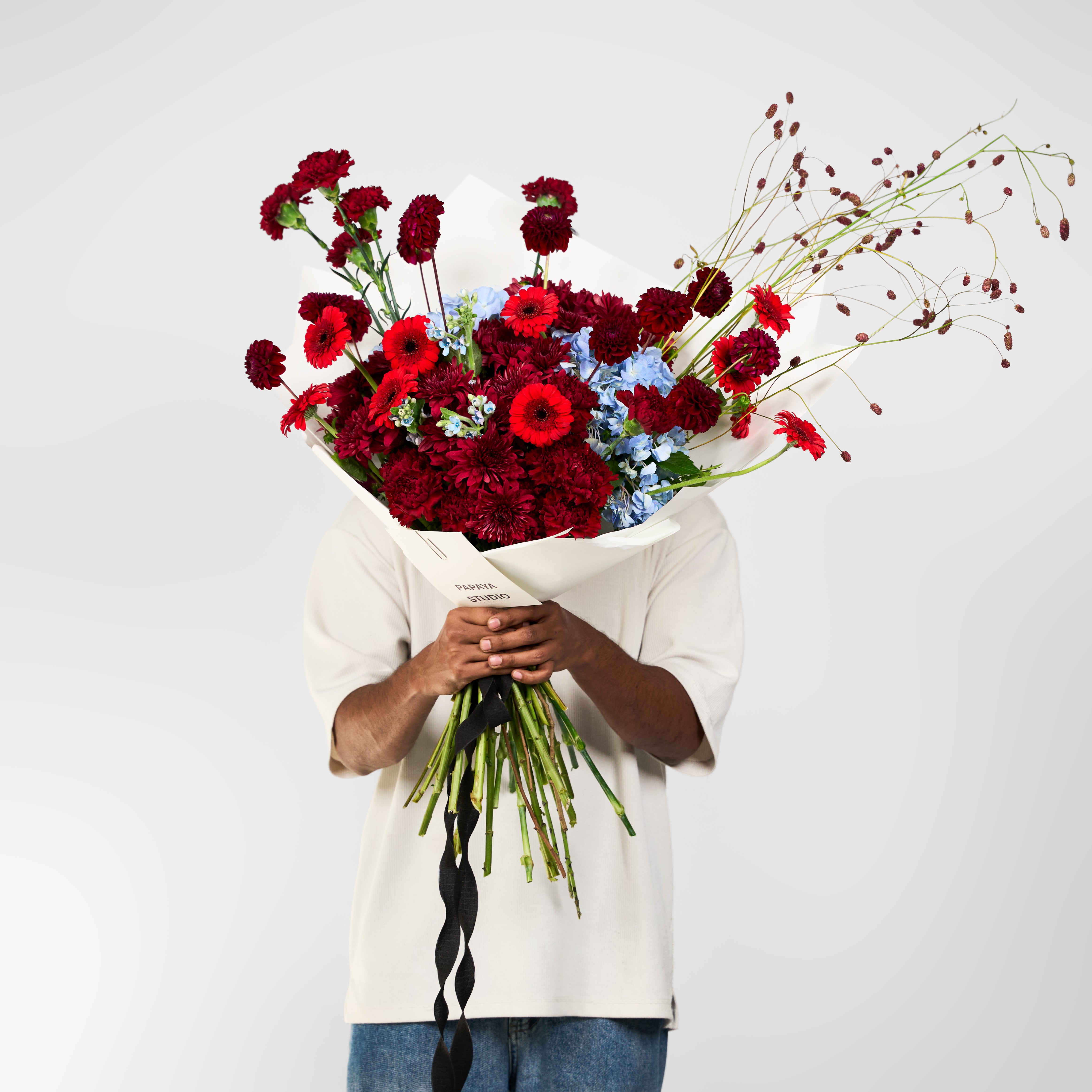 Red and burgundy bouquet with pale blue flowers, airy branching stems, ivory wrapping and a black ribbon